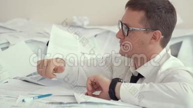 一个戴眼镜的疲惫的年轻人坐在一堆文件和签名文件里。 因为疲劳，一个人心烦意乱